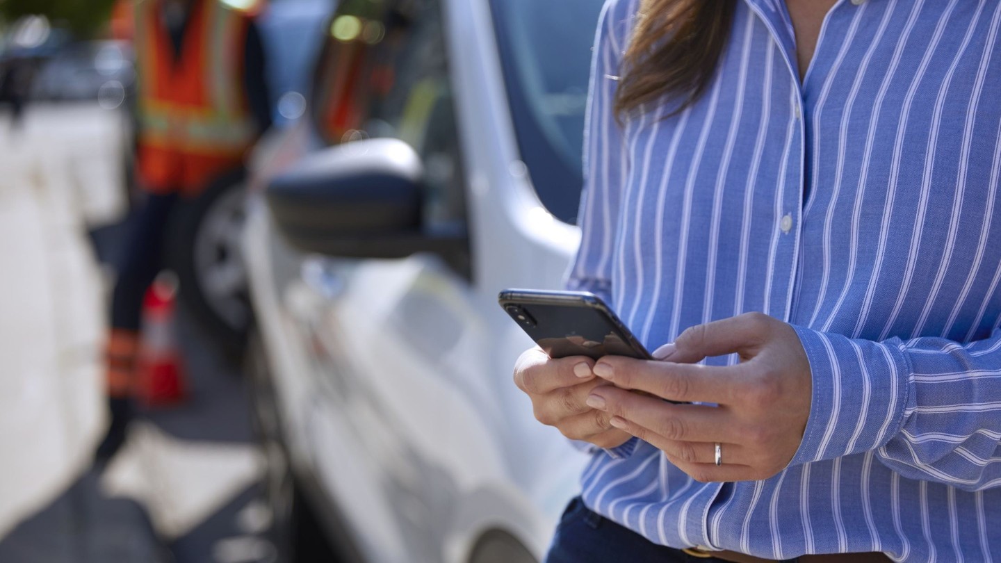 Una mujer en primer plano usando un smartphone, apoyada en un coche y un operario con un chaleco de alta visibilidad al fondo.