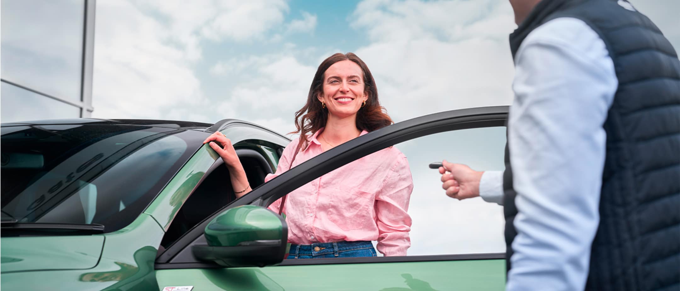 Una mujer sonriente con camisa rosa y vaqueros está junto a la puerta abierta de un coche verde, recibiendo la llave del coche de un hombre