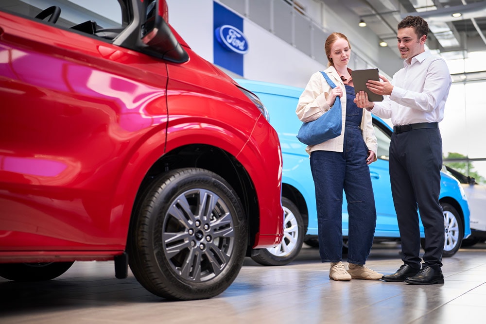 Dos personas en un concesionario Ford mirando una tableta, con una furgoneta Ford roja en primer plano y una furgoneta Ford azul detrás.