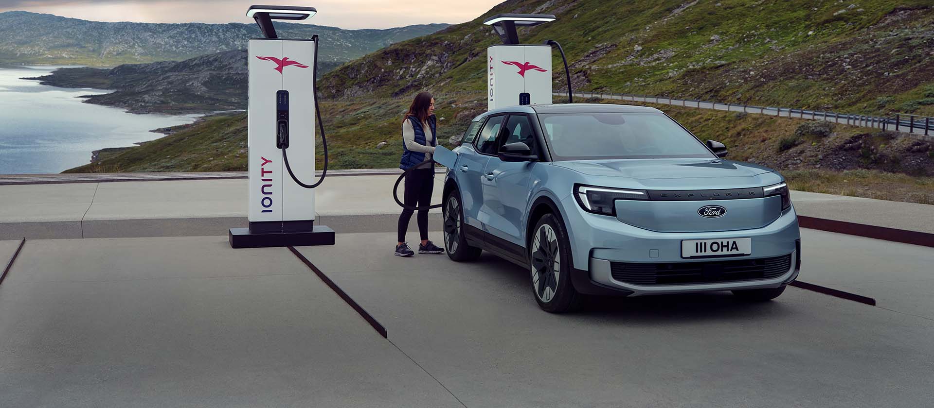 Una mujer conecta un Ford Explorer azul, totalmente eléctrico, a una estación de carga pública usando un cable. El coche está aparcado cerca de montañas y agua.