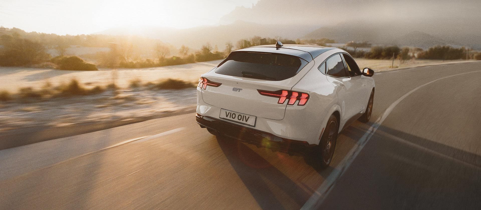 Un vehículo eléctrico Ford Mustang Mach-E plateado conduciendo por una carretera larga y abierta.