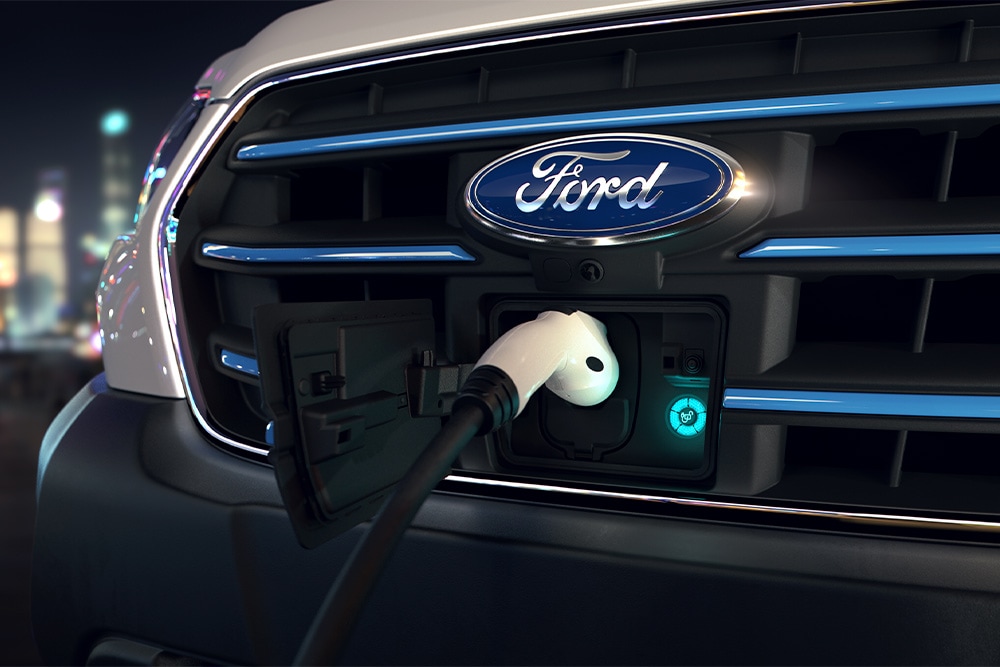Primer plano de un punto de carga de una furgoneta Ford E-Transit y la insignia ovalada azul de Ford en la parrilla delantera.