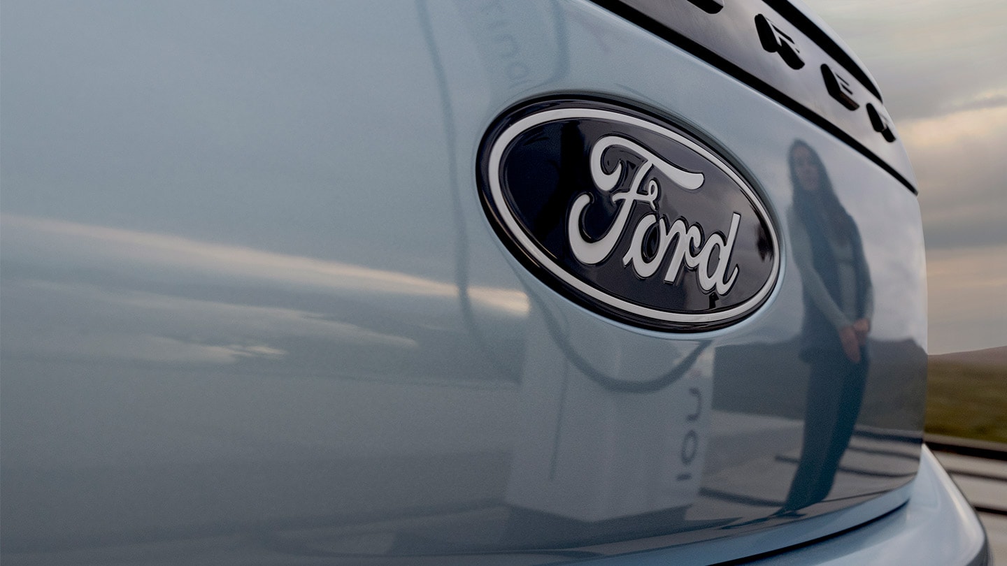 Logotipo de Ford en un Ford Explorer eléctrico azul con una estación de carga pública reflejada en el automóvil.