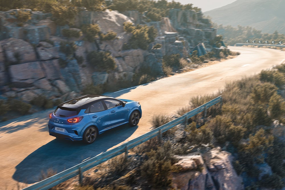 Imagen de un Puma Gen-E azul circulando por una sinuosa carretera de montaña.