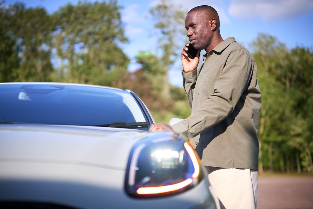 Una persona al teléfono junto a un coche de color claro al aire libre. 