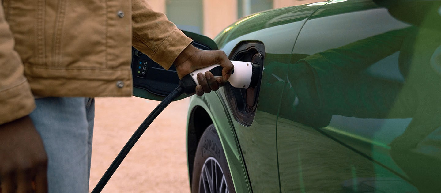 Una persona con una chaqueta color crema conecta un cable de carga eléctrica a su Ford Mustang Mach-E en Green Electric.