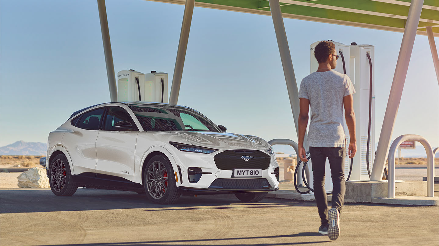 Un Ford Mustang Mach-E blanco estacionado en una estación de carga pública exterior. Un hombre con una camiseta blanca caminando hacia un coche.