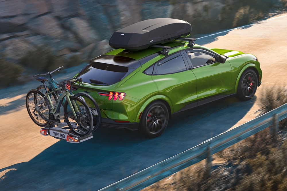 Vista trasera de un Ford Mustang Mach-E verde, equipado con un cofre de techo y un portabicicletas, circulando por una carretera panorámica. 