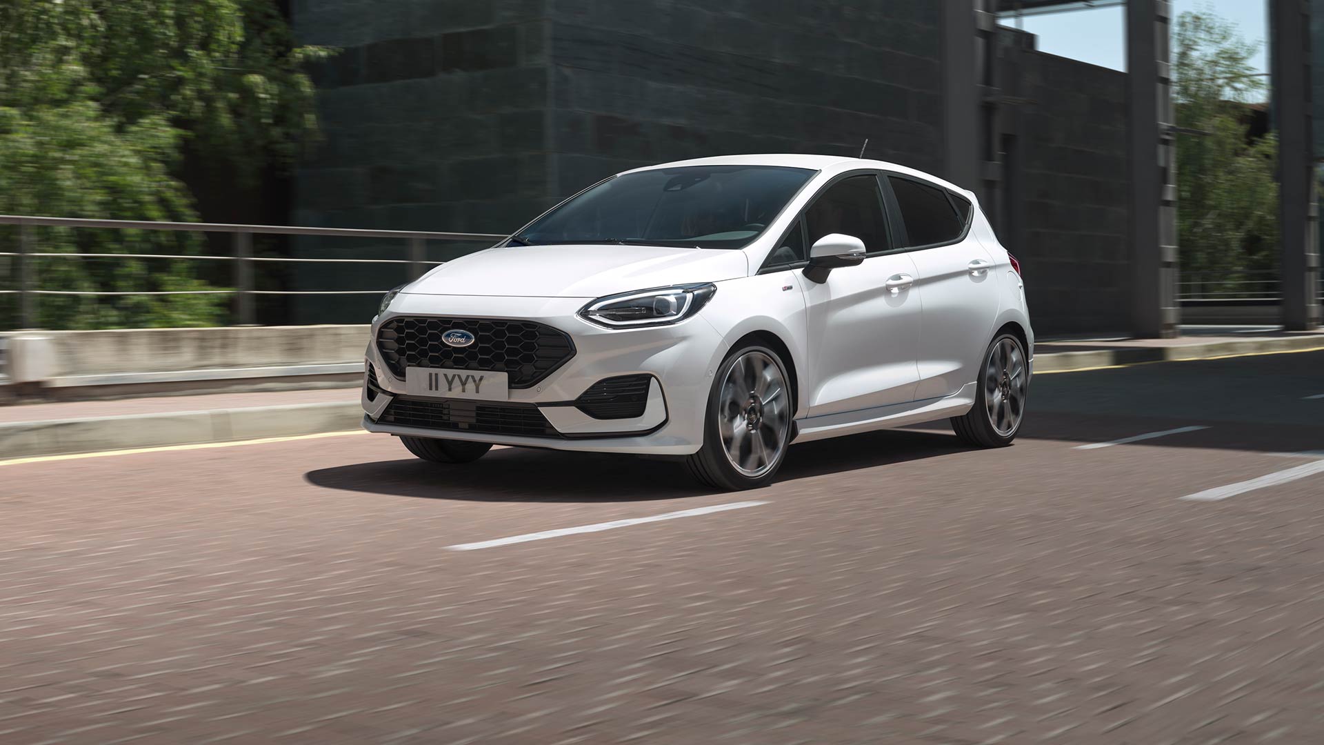 Un Ford Fiesta blanco circulando por una calle de ciudad, con zonas verdes y edificios al fondo.