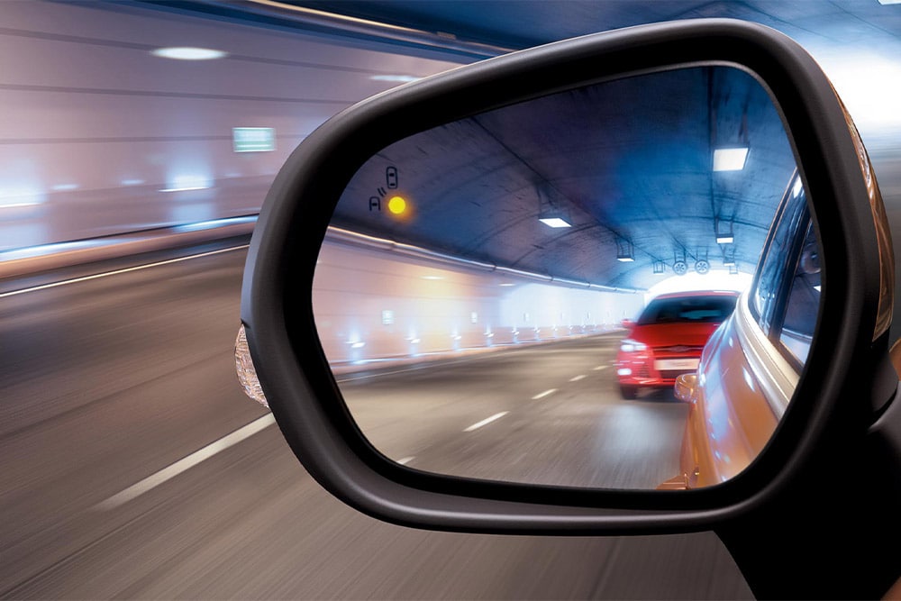 Un primer plano del espejo retrovisor lateral en un túnel, que muestra un automóvil rojo que se acerca en el punto ciego del conductor.