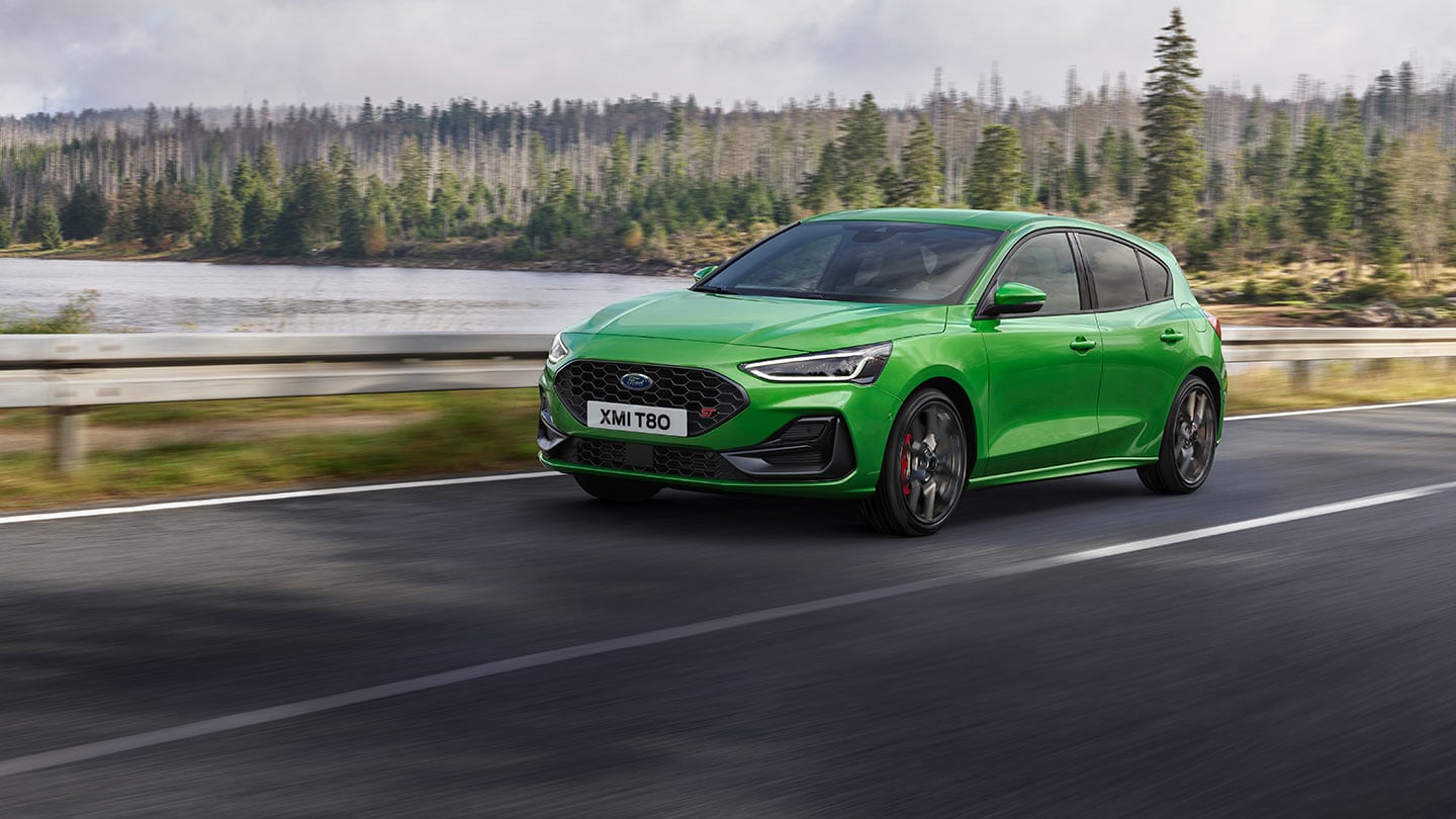 Un Ford Focus ST en Mean Green conduciendo por una carretera con árboles al fondo.