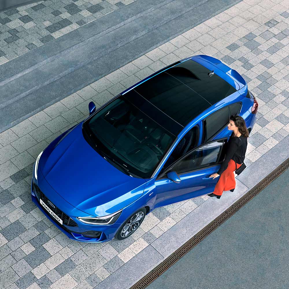 Una vista aérea de una mujer subiendo a su Ford Focus azul que tiene un techo panorámico de vidrio.