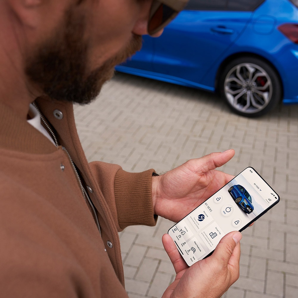 Una persona sosteniendo un teléfono inteligente que muestra la aplicación FordPass™, con un Ford Focus azul en el fondo.