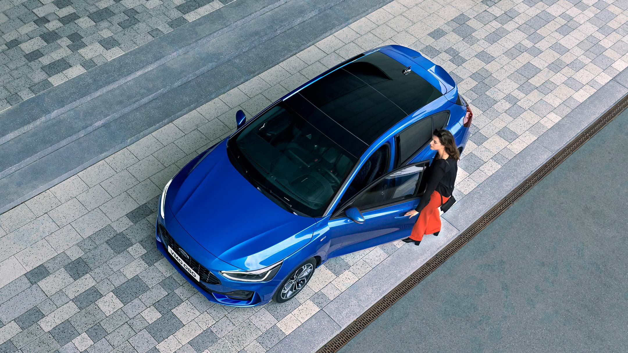 Una vista aérea de una mujer subiendo a su Ford Focus azul que tiene un techo panorámico de vidrio.