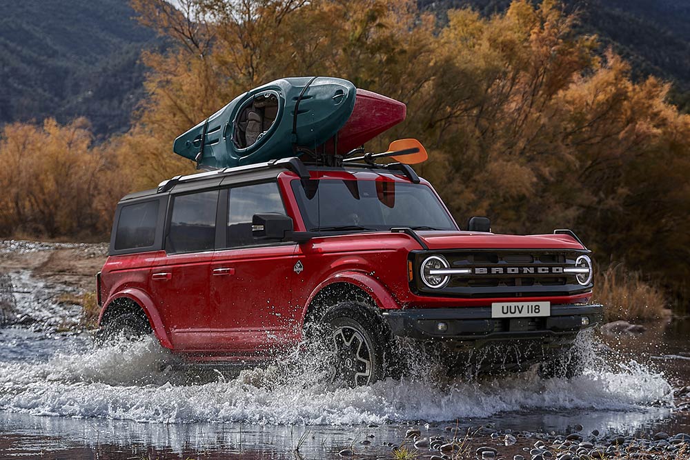 Vista lateral de un Ford Bronco rojo atravesando aguas poco profundas en un paisaje montañoso.