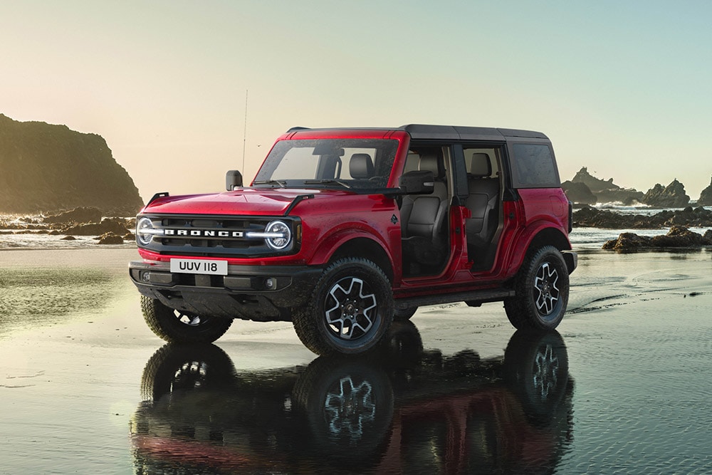 Vista lateral de un Ford Bronco rojo, con las puertas desmontadas, aparcado en una orilla arenosa.