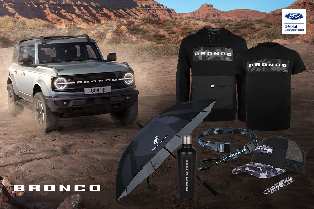 Ford Bronco Collection with black t-shirt, jacket, polo, cap, beanie, and wetpack, and a grey Bronco in the background.