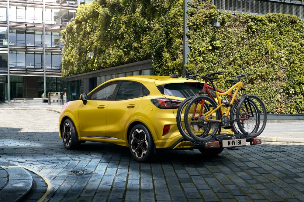 Ford Puma Gen-E amarillo aparcado en un entorno urbano con dos bicicletas montadas en un portabicicletas trasero.