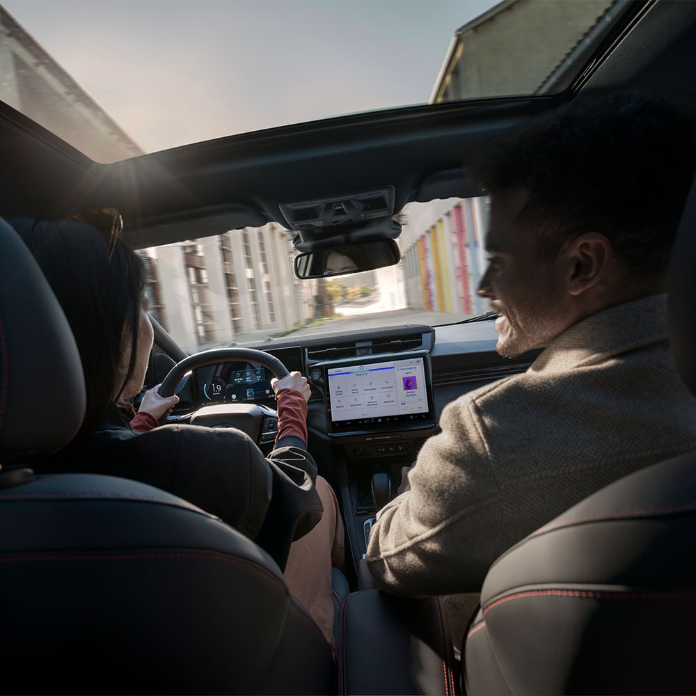 Una persona conduciendo conversa con la que va en el asiento del pasajero mientras el vehículo recorre un paisaje residencial soleado.