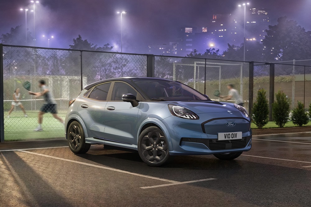 Un Ford Puma Gen-E azul aparcado junto a una pista de tenis, con las luces de la ciudad al fondo.