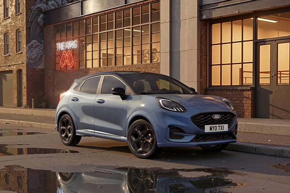 Un Ford Puma azul aparcado en la calle de una ciudad al atardecer, con charcos en los que se reflejan los edificios.