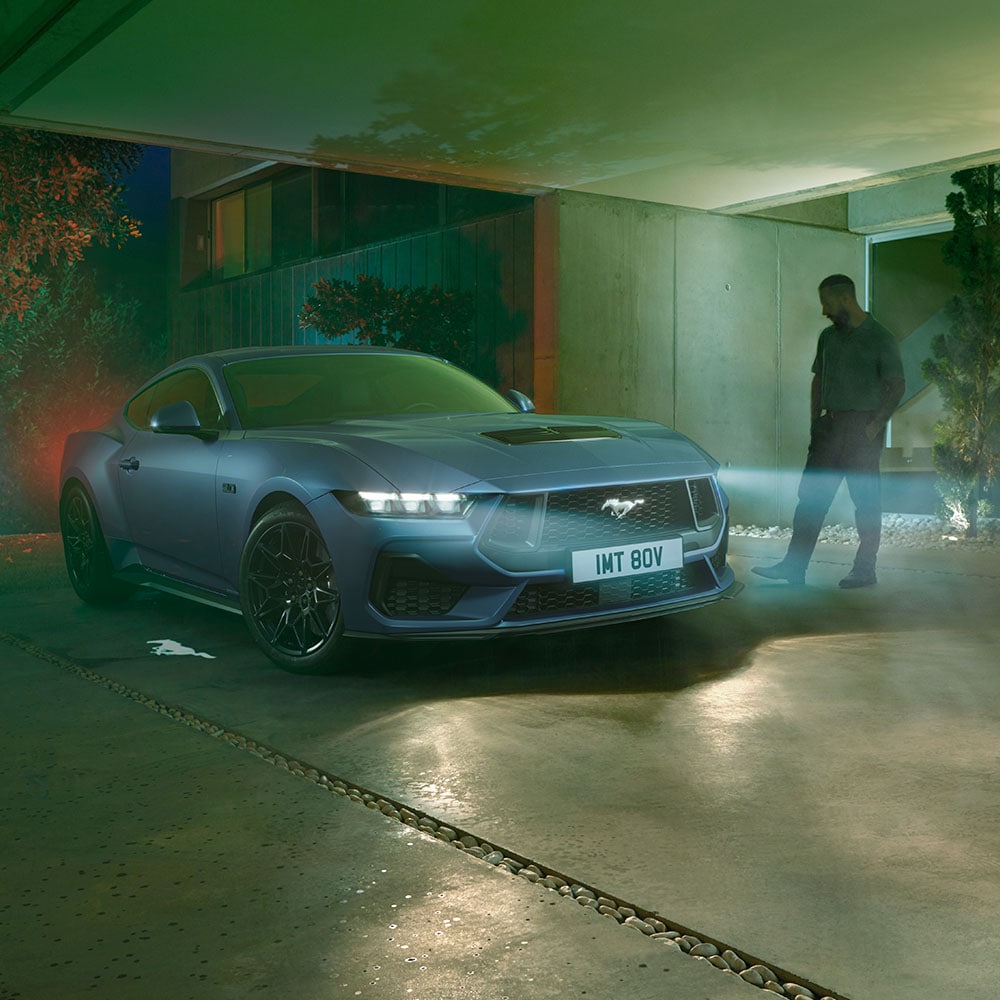 Hombre acercándose a un Ford Mustang estacionado con los faros encendidos, mostrando la iluminación de bienvenida al detectar la aproximación