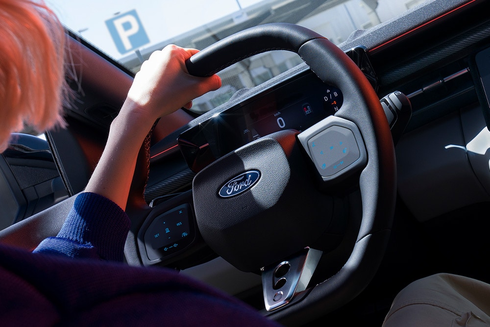 Primer plano de una mujer con las manos sobre el volante de un Ford Capri estacionado, donde puede verse asimismo el cuadro de instrumentos digital del vehículo.