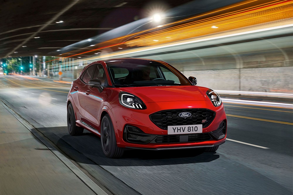 Un SUV deportivo Ford Puma ST rojo circula por una carretera urbana iluminada por la noche.