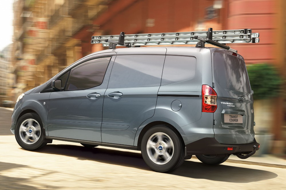 Ford Transit Courier de color azul plata parada en la carretera con portaequipajes y escalera. Vista de tres cuartos trasera