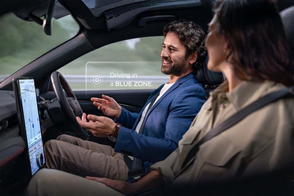 Un hombre y una mujer en un Mustang Mach-E conduciendo sin manos con Ford BlueCruise. Una notificación dice "Conduciendo en una BLUE ZONE".