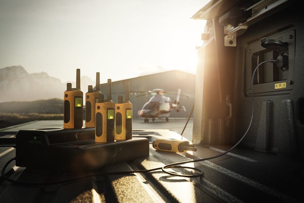 Varios walkie-talkies cargándose en la plataforma de carga de un Ranger Raptor, con un helicóptero de fondo, al atardecer.