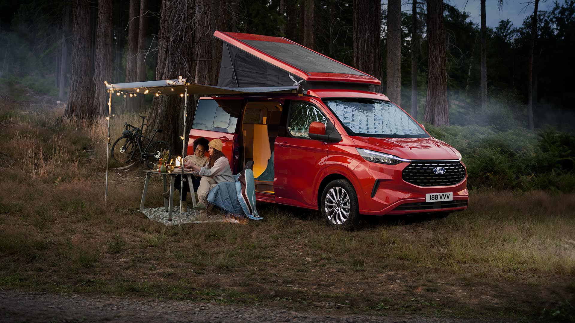 Dos mujeres sentadas debajo de un toldo que sobresale de las puertas laterales abiertas de una Ford Transit Custom Nugget roja, con el techo inclinado hacia arriba.