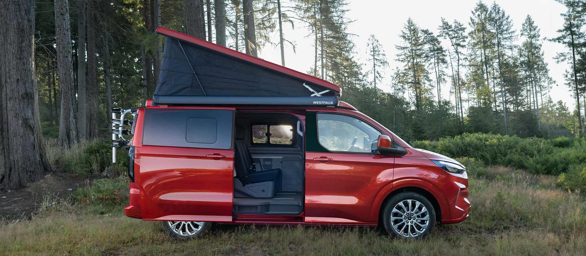 Ford Transit Custom Nugget roja aparcada con el techo con tejido autoplegable inclinado hacia arriba y la puerta lateral deslizante abierta.