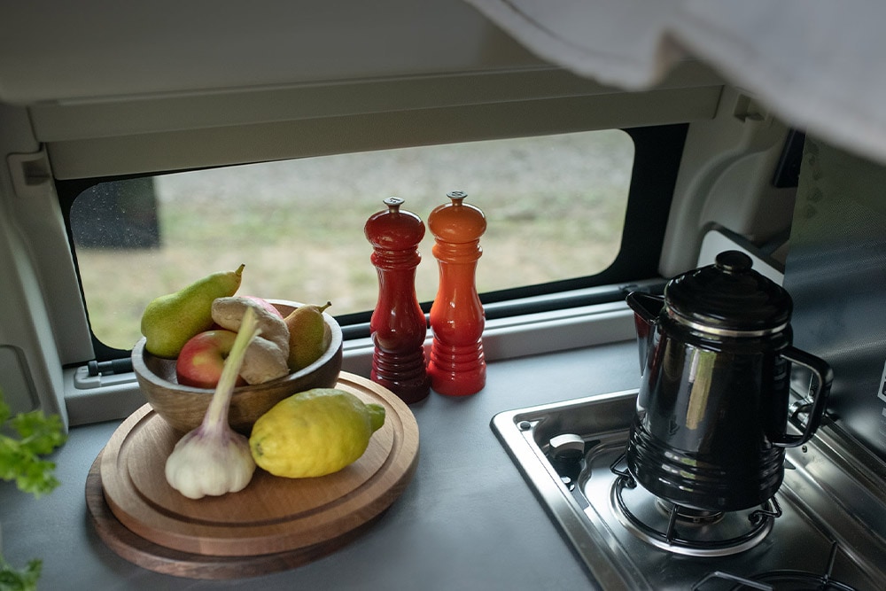 Fogón de la cocina integrada de la Ford Transit Custom Nugget, con un hervidor y fruta en la encimera.