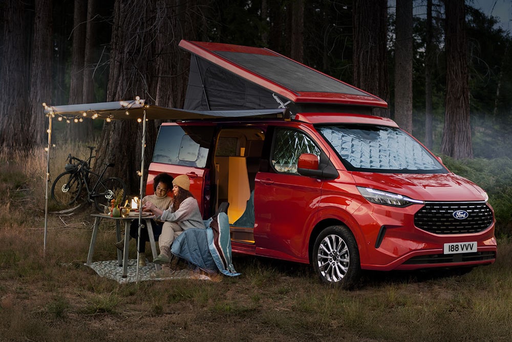 Dos mujeres sentadas debajo de un toldo que sobresale de las puertas laterales abiertas de una Ford Transit Custom Nugget roja, con el techo inclinado hacia arriba.