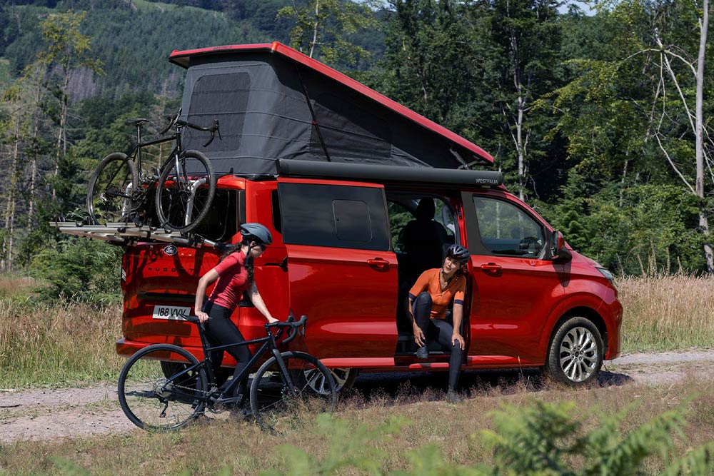 Personas descargando las bicicletas de un portabicicletas de una Ford Transit Custom Nugget roja.