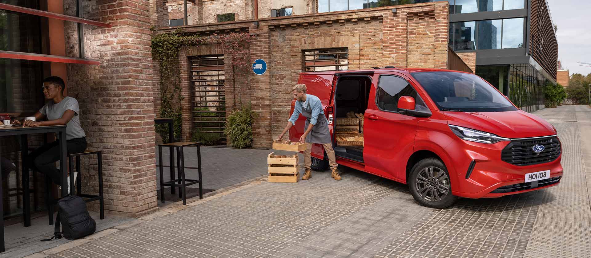 Una persona descarga cajas de una furgoneta Ford Transit Custom roja estacionada frente a una cafetería. 