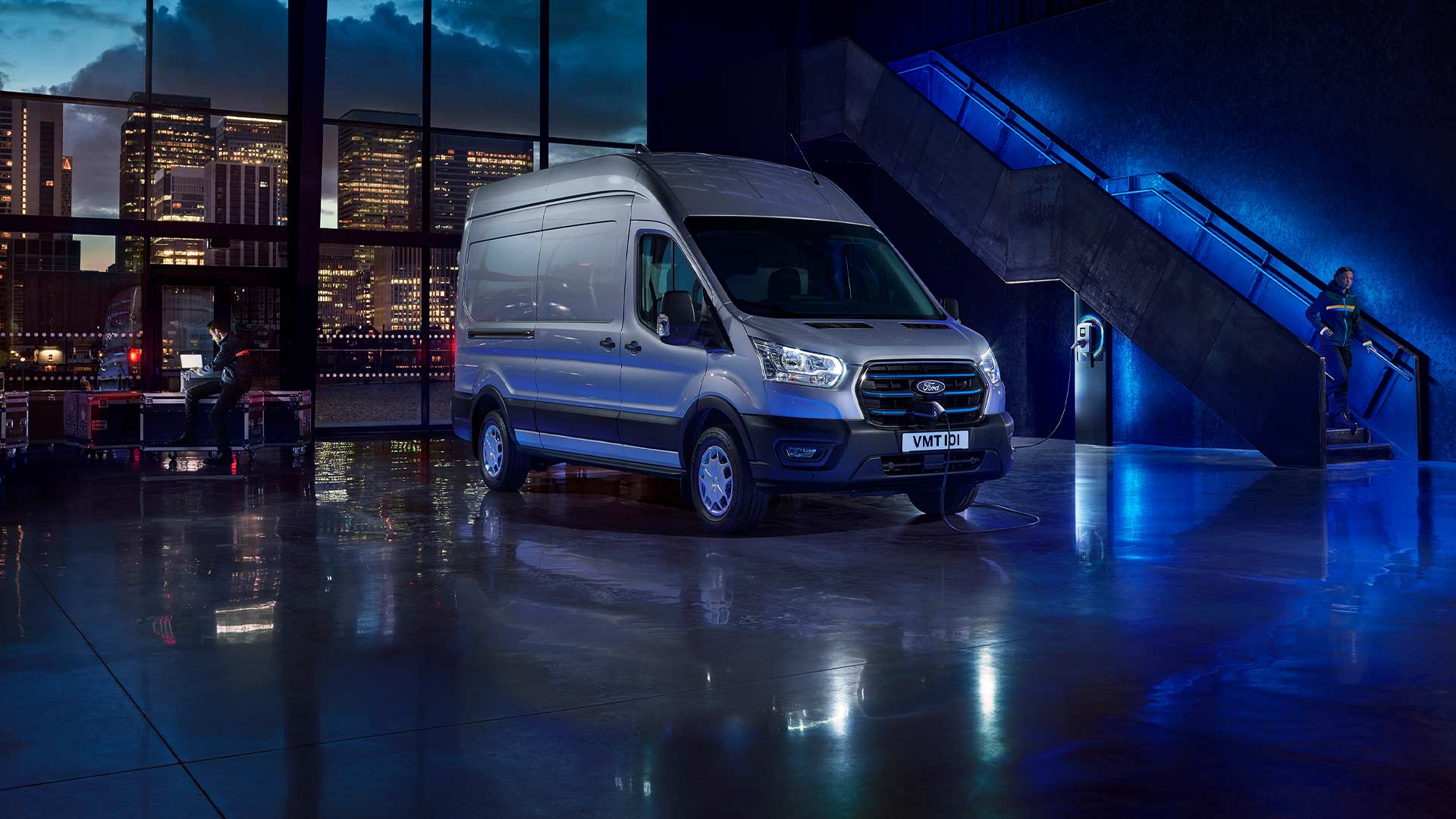 Furgoneta eléctrica Ford E-Transit en color plateado aparcada en el interior de un recinto desde el que se ve la línea del horizonte de una ciudad a través de grandes ventanales.