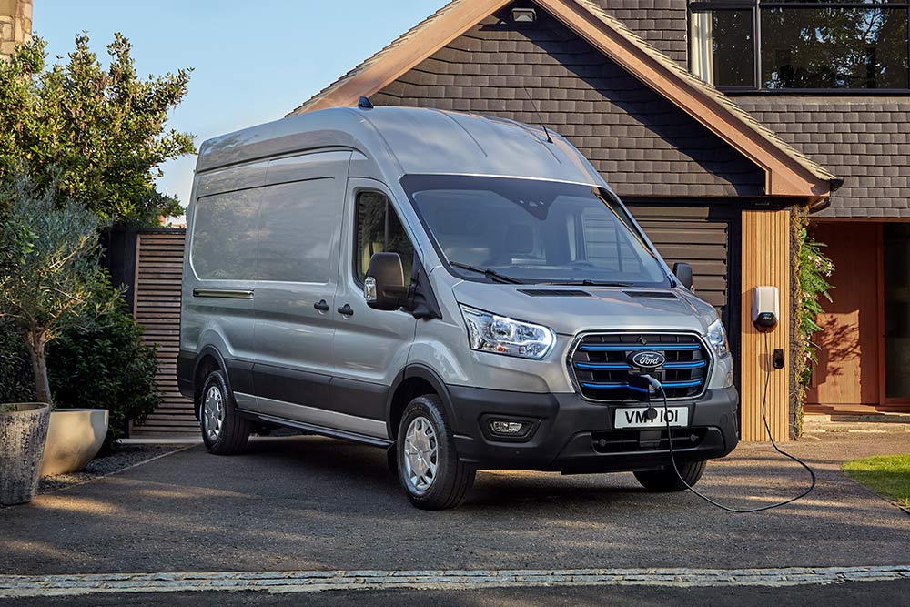 Furgoneta Ford E-Transit plateada aparcada en un camino de entrada de una casa, conectada a una estación de carga de vehículos eléctricos.