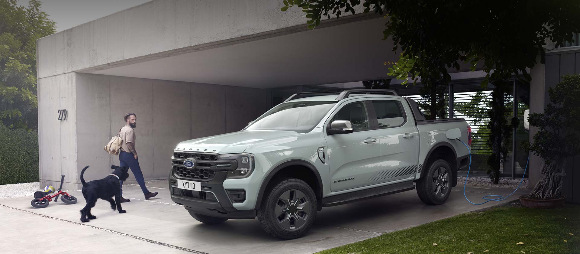 Un hombre y su perro entran en una casa moderna, pasando al lado de un Ford Ranger híbrido enchufable Stormtrak que se encuentra cargando. 