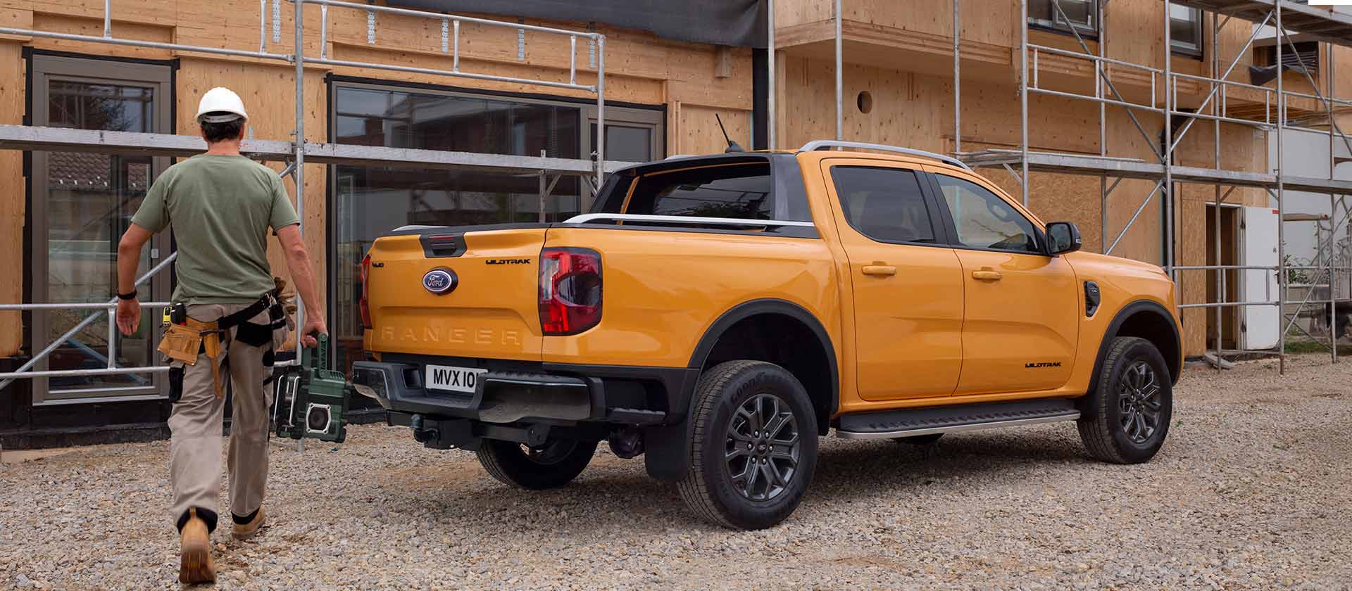Un trabajador con casco camina hacia un modelo pick-up Ford Ranger amarillo estacionado en una parcela en construcción.