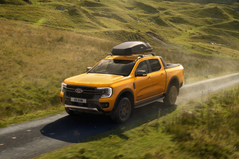 Ford Ranger amarillo circulando por una carretera rural con una caja portaequipajes montada sobre las barras transversales del techo.