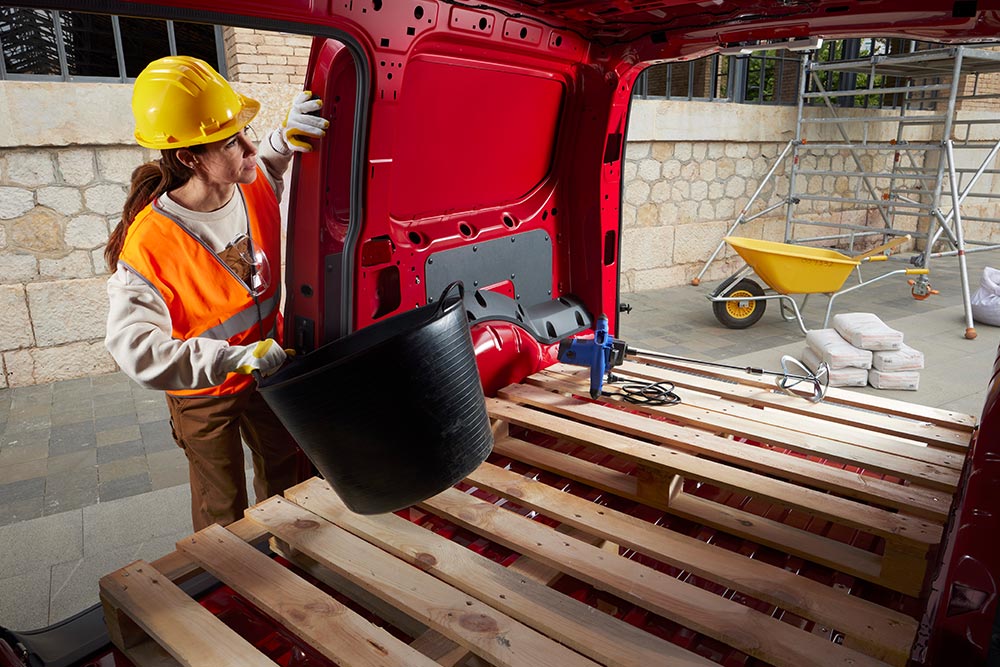 Una mujer coloca materiales de construcción en el espacio de carga de una Ford Transit Connect con la puerta lateral y las traseras abiertas.