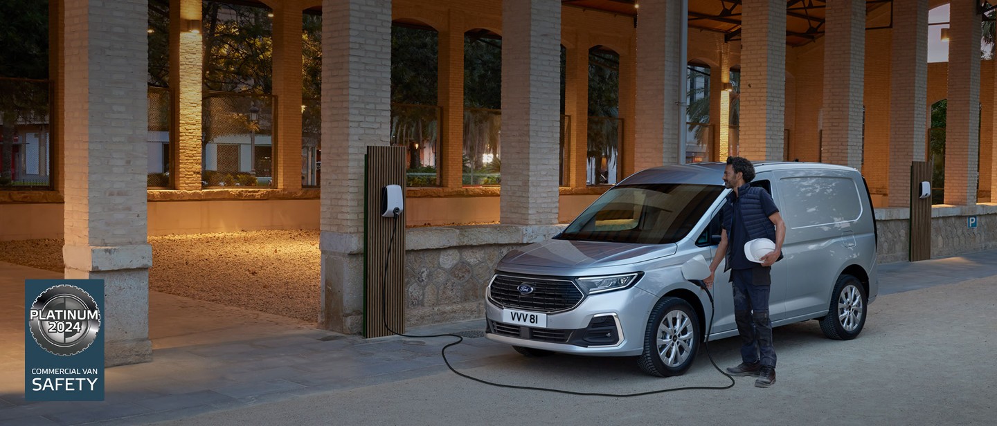 Un hombre que sostiene un casco enchufa un cable de carga a la caja de pared en la nueva Ford Transit Connect en Stardust Silver.