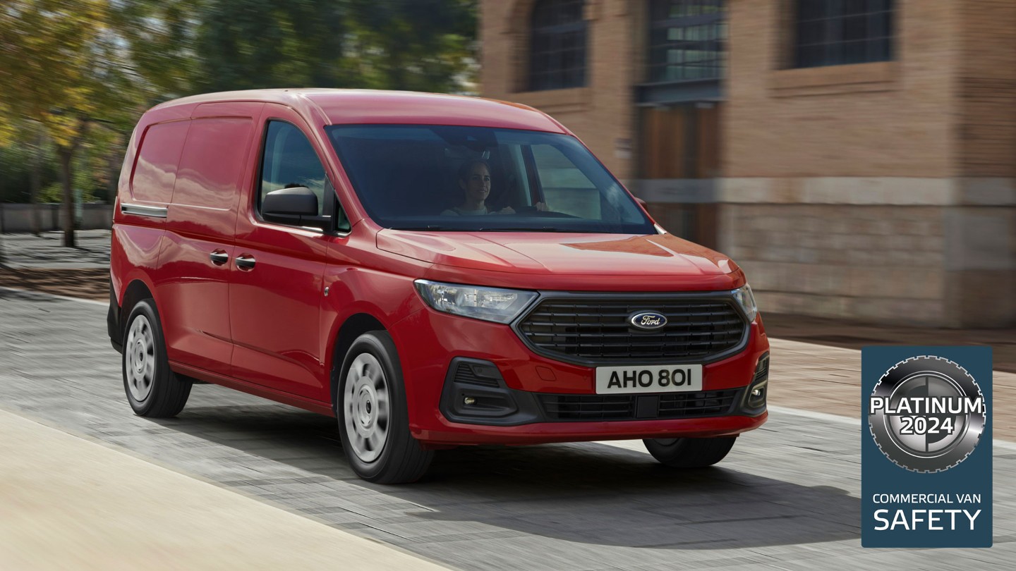 La nueva Ford Transit Connect Trend en color rojo arce conduce por una calle y muestra la parrilla delantera y los faros automáticos. 