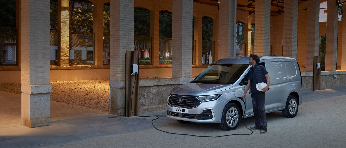 Un hombre que sostiene un casco enchufa un cable de carga a la caja de pared en la nueva Ford Transit Connect en Stardust Silver.
