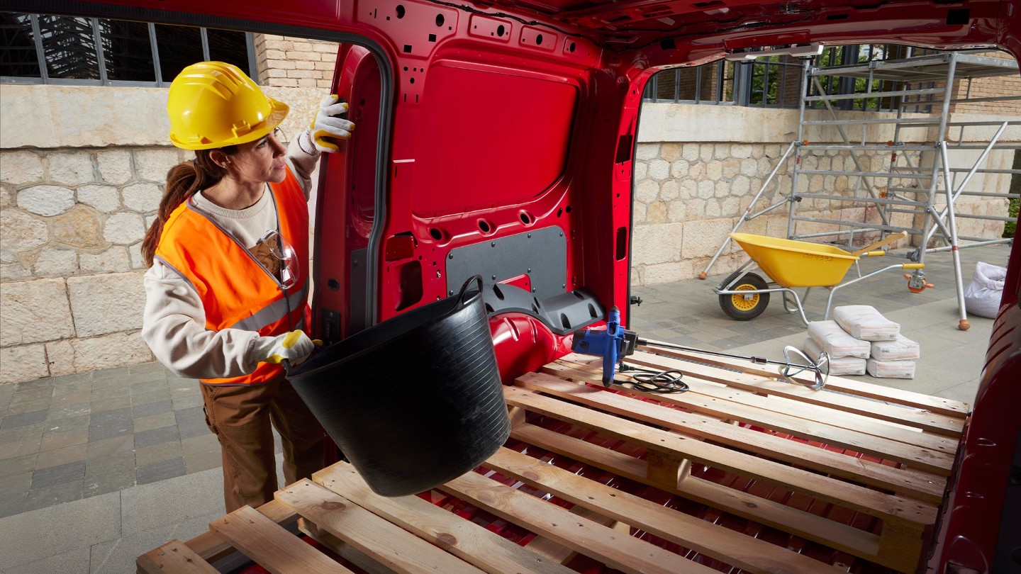 Una mujer coloca materiales en el espacio de carga de la nueva Ford Transit Connect en rojo arce con las puertas laterales y traseras abiertas, en una obra de construcción. 