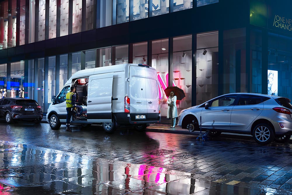 Una Ford Transit blanca con la puerta lateral abierta estacionada entre dos coches en una vía urbana lluviosa. 