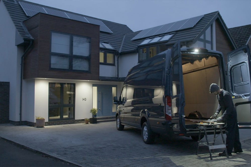 Un carpintero trabaja detrás de una furgoneta Ford Transit con las puertas traseras abiertas por la noche, iluminando el espacio de trabajo con la luminaria LED trasera integrada.