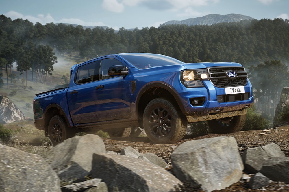 Una pick-up Ford Ranger azul circula por un camino de tierra en un bosque, dejando un rastro de polvo.